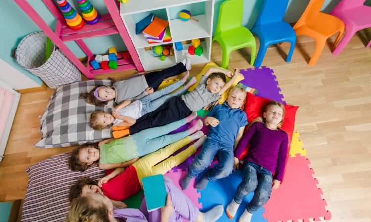 Young attractive teacher reading story before the naptime at the kindergarten. Lovely kids laying on a floor on soft mats and pillows. Time to have a rest and to calm down. Top view.